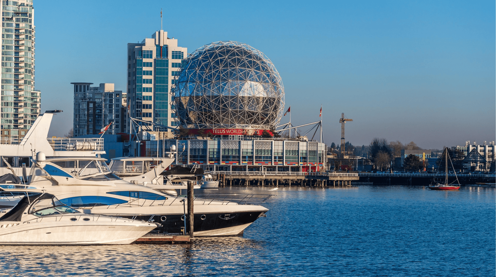 Boats docked in False Creek with the TELUS World of Science geodesic dome and modern Vancouver skyline in the background on a clear blue day