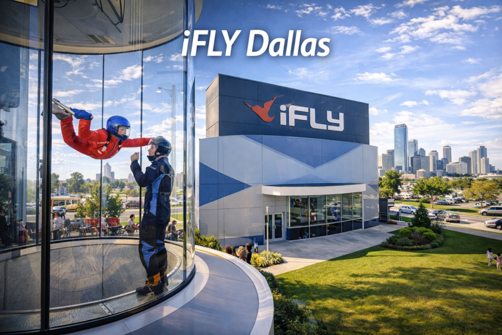 iFLY Dallas indoor skydiving wind tunnel with instructor guiding a first time flyer and Dallas skyline visible under bright daylight.